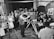 A vintage television studio setting with a large group of people gathered around a cameraman operating an old-fashioned television camera. The audience is dressed in formal attire typical of the mid-20th century. The studio has a brick wall backdrop with cartoonish artwork.