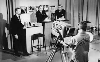 A vintage television studio scene with three men in suits engaging in a discussion or interview, while a cameraman operates a large, old-fashioned TV camera. In the background, there are musical records displayed on a shelf, and a drum set is visible to the side.