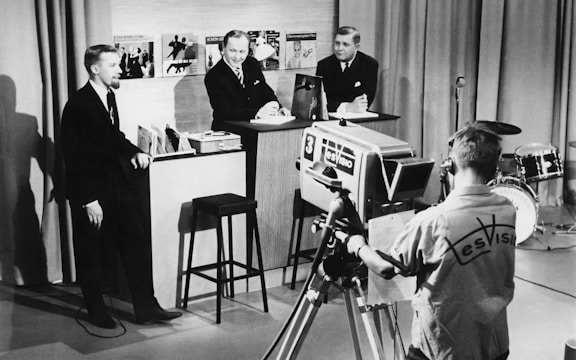 A vintage television studio scene with three men in suits engaging in a discussion or interview, while a cameraman operates a large, old-fashioned TV camera. In the background, there are musical records displayed on a shelf, and a drum set is visible to the side.