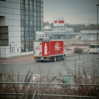 A Coca-Cola delivery truck is parked near a modern building with large glass windows. The truck is prominently branded with the Coca-Cola logo and bottle design. Several other vehicles are visible, and the scene includes bare shrubbery and a road sign indicating a no-parking zone. The background features more buildings and a billboard, with a cloudy sky and muted light suggesting an overcast day.