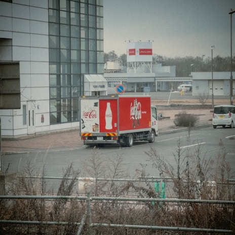 A Coca-Cola delivery truck is parked near a modern building with large glass windows. The truck is prominently branded with the Coca-Cola logo and bottle design. Several other vehicles are visible, and the scene includes bare shrubbery and a road sign indicating a no-parking zone. The background features more buildings and a billboard, with a cloudy sky and muted light suggesting an overcast day.