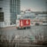 A Coca-Cola delivery truck is parked near a modern building with large glass windows. The truck is prominently branded with the Coca-Cola logo and bottle design. Several other vehicles are visible, and the scene includes bare shrubbery and a road sign indicating a no-parking zone. The background features more buildings and a billboard, with a cloudy sky and muted light suggesting an overcast day.