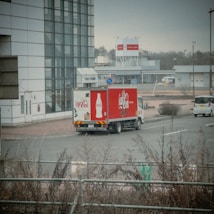 A Coca-Cola delivery truck is parked near a modern building with large glass windows. The truck is prominently branded with the Coca-Cola logo and bottle design. Several other vehicles are visible, and the scene includes bare shrubbery and a road sign indicating a no-parking zone. The background features more buildings and a billboard, with a cloudy sky and muted light suggesting an overcast day.