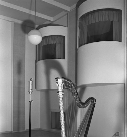 A vintage-style black and white photograph depicting a harp leaned against a wall in a possibly soundproofed room. A microphone stands on a stand next to it while two uniquely designed windowed rooms in the background are visible, complete with curtains. The shadows of the harp and microphone are cast on the wall, adding depth to the image.