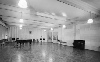 A large, empty room with a high ceiling, illuminated by several hanging lights. The room has wooden flooring and a combination of paneled and plain walls. A grand piano and upright piano are present, along with rows of empty chairs and a few music stands positioned near the walls. There is a microphone stand in the middle of the room and some large windows with blinds on the far left.