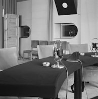 A vintage recording studio features a table with microphones arranged in a row. The room includes padded chairs, a wooden door on the left, and a window with a viewing port. Overhead, a directional light hangs down, and there is minimal decor with plain walls.