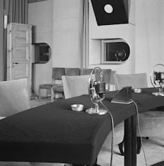 A vintage recording studio features a table with microphones arranged in a row. The room includes padded chairs, a wooden door on the left, and a window with a viewing port. Overhead, a directional light hangs down, and there is minimal decor with plain walls.