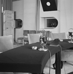A vintage recording studio features a table with microphones arranged in a row. The room includes padded chairs, a wooden door on the left, and a window with a viewing port. Overhead, a directional light hangs down, and there is minimal decor with plain walls.