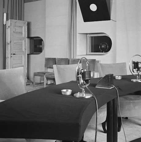 A vintage recording studio features a table with microphones arranged in a row. The room includes padded chairs, a wooden door on the left, and a window with a viewing port. Overhead, a directional light hangs down, and there is minimal decor with plain walls.