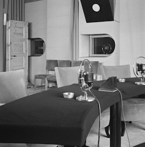A vintage recording studio features a table with microphones arranged in a row. The room includes padded chairs, a wooden door on the left, and a window with a viewing port. Overhead, a directional light hangs down, and there is minimal decor with plain walls.