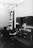 A man is seated at a desk filled with various radio equipment and controls in a room that appears to be an old-fashioned radio or communication station. The equipment includes dials, gauges, and switches along the walls, with a focus on the central console he is operating.