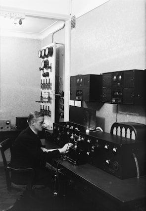 A man is seated at a desk filled with various radio equipment and controls in a room that appears to be an old-fashioned radio or communication station. The equipment includes dials, gauges, and switches along the walls, with a focus on the central console he is operating.