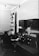 A man is seated at a desk filled with various radio equipment and controls in a room that appears to be an old-fashioned radio or communication station. The equipment includes dials, gauges, and switches along the walls, with a focus on the central console he is operating.