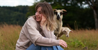 A woman sits in a grassy field, smiling and resting her head on her hand. Behind her, a sheep and a lamb stand, enhancing the pastoral setting. The background features dense, green foliage.