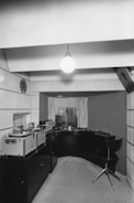 A vintage audio studio setup featuring large reel-to-reel tape machines on a counter to the left. The room has a minimalist design with wood paneling and a single hanging light bulb. A swivel chair is positioned in front of a curved desk space that houses more audio equipment, creating an intimate and focused work area. The window in the background is partially covered with curtains, allowing some light to filter through.