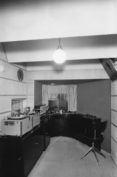 A vintage audio studio setup featuring large reel-to-reel tape machines on a counter to the left. The room has a minimalist design with wood paneling and a single hanging light bulb. A swivel chair is positioned in front of a curved desk space that houses more audio equipment, creating an intimate and focused work area. The window in the background is partially covered with curtains, allowing some light to filter through.