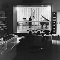 A black and white photograph of a recording studio with a large window separating the control room from the studio space. Inside the studio, a grand piano and music stands are visible. A person stands in front of a large wall clock, facing away from the camera. In the foreground, the control room features vintage phones and an array of control panels with buttons and switches.
