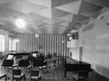 An acoustic room with a geometric, patterned ceiling, featuring a grand piano, several chairs in various arrangements, and music stands. The room has a bright atmosphere due to the large windows on the left, and round pendant lights hanging from the ceiling add to the lighting.