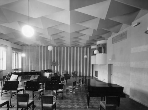An acoustic room with a geometric, patterned ceiling, featuring a grand piano, several chairs in various arrangements, and music stands. The room has a bright atmosphere due to the large windows on the left, and round pendant lights hanging from the ceiling add to the lighting.