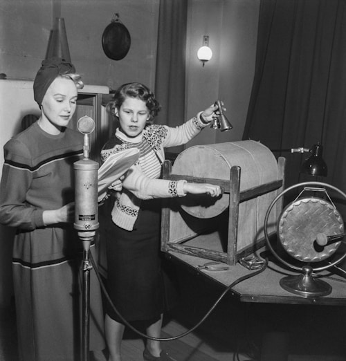 Two women are in a room engaged in a radio broadcast setting. One woman is speaking into a vintage microphone while holding papers, suggesting she is reading or announcing. The other woman is operating a sound effects device, possibly hand-cranked, to create audio effects. The scene has a classic mid-20th-century appearance with period clothing and broadcasting equipment.