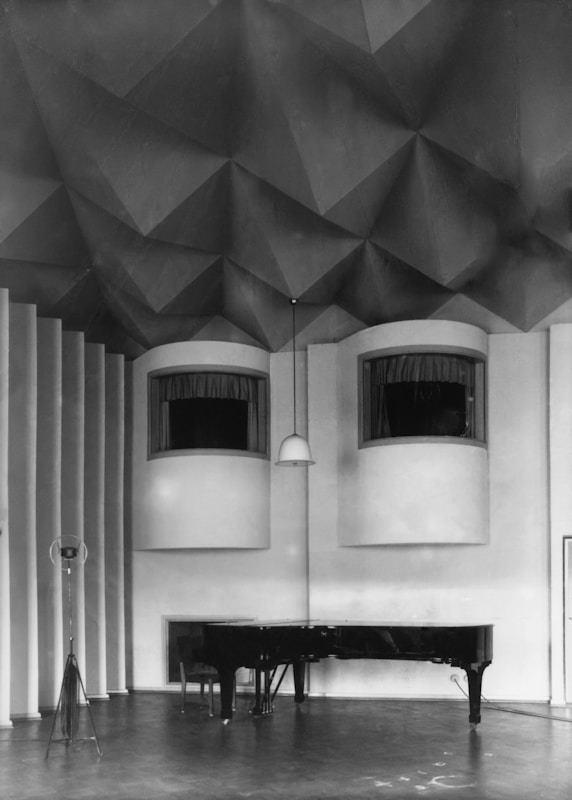 A black grand piano is positioned in the corner of a geometric, acoustically designed room. The ceiling features an intricate pattern of three-dimensional, faceted shapes. Two modern half-circle windows are set into a textured wall, and a hanging light fixture is centered between them. The floor is smooth with a slightly glossy finish, and a microphone stand is visible on the left side of the room.