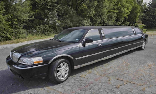 A long, black limousine is parked on a cracked asphalt surface with lush green trees in the background. The vehicle features a sleek design with silver rims and tinted windows.