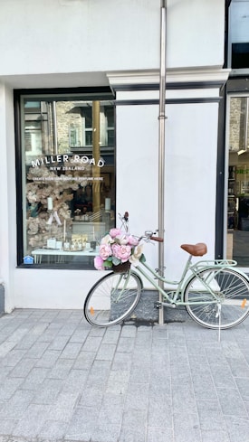 A vintage-style bicycle with a brown leather seat and a front basket filled with pink roses is parked on a paved sidewalk. Behind the bicycle, there is a shop window displaying perfume bottles and floral decorations. The window has the text 'Miller Road New Zealand' indicating the store's name.