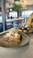 A cozy cafe interior with wooden stools and a chandelier. A large plate displays a dessert with bread topped with cream and banana slices, while another dish features a savory toast with garnish.