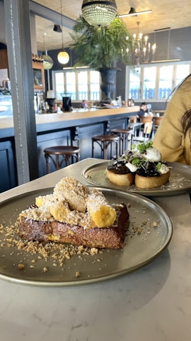 A cozy cafe interior with wooden stools and a chandelier. A large plate displays a dessert with bread topped with cream and banana slices, while another dish features a savory toast with garnish.