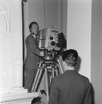 A man in a suit is operating a large, vintage television camera mounted on a tripod. He is wearing a headset with a microphone. Another man, also in a suit, is standing with his back to the camera, observing. The setting appears to be indoors, with part of an architectural structure visible on the left.
