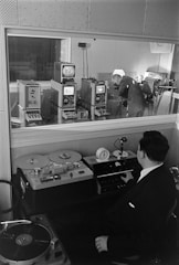 A man in a suit sits at a console with various audio equipment, including a reel-to-reel tape recorder and a turntable. Through a glass partition, another man in business attire is seen leaning over electronic devices, suggesting an audio or broadcast studio setting.