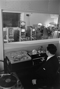 A man in a suit sits at a console with various audio equipment, including a reel-to-reel tape recorder and a turntable. Through a glass partition, another man in business attire is seen leaning over electronic devices, suggesting an audio or broadcast studio setting.