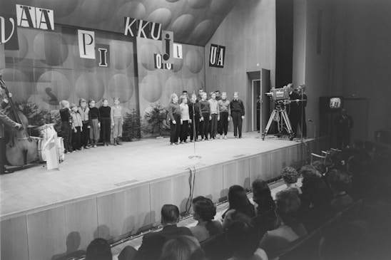 A black and white photograph of a stage with a group of children in formal attire, standing in a line and possibly singing or performing. A camera crew is visible, recording the event. The audience is seated in the foreground, watching attentively. The background includes large, decorative letters and a textured wall.