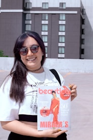 A person smiles broadly while holding a magazine in front of a modern building with multiple windows. They wear sunglasses, a white t-shirt with text, a black choker, and black overall straps. The magazine has a cover featuring a model in a red dress.