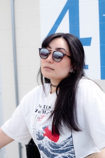 A person with long dark hair wearing sunglasses and a white graphic t-shirt stands against a wall with blue lettering. The t-shirt features a design with waves and a red circle, reminiscent of Japanese art.