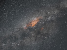 A dense cluster of stars and cosmic dust is vividly portrayed against a vast, dark expanse. The central region features a vibrant concentration of orange and yellow hues, suggesting areas rich in stellar activity. Surrounding this bright core, the space is filled with a plethora of tiny luminous dots representing distant stars, contributing to the grandeur and depth of the celestial scene.