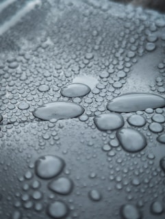Numerous droplets of water varying in size cover a smooth surface, creating a pattern of circles that seem to overlap each other. The droplets have a reflective quality, contributing to a glossy appearance.