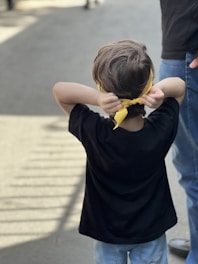 A child stands outdoors with their back to the camera, tying a yellow bandana around their head. They are wearing a black shirt and light blue jeans. The setting appears to be a sunny day with shadows visible on the pavement. An adult stands nearby wearing blue jeans.