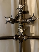 A vintage metal shower valve with four handles, each labeled with a different function: Hot, Cold, Shower, and Needle. The central structure is crafted from metal pipes with a slightly tarnished appearance, and the handles have a classic design.