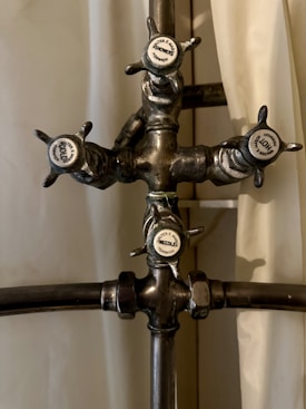A vintage metal shower valve with four handles, each labeled with a different function: Hot, Cold, Shower, and Needle. The central structure is crafted from metal pipes with a slightly tarnished appearance, and the handles have a classic design.