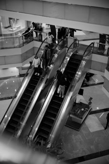 A black and white image of an indoor shopping mall featuring two escalators. Several people are using the escalators, some ascending and others descending. The surrounding area includes glass railings, store displays, and interior decor elements.