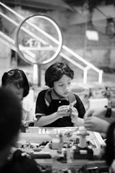 Children are sitting at a table engaged in building with LEGO bricks. The background features a large circular light fixture and a bustling indoor environment, suggesting a playful or educational setting.