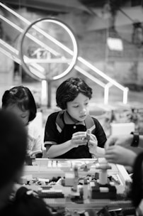 Children are sitting at a table engaged in building with LEGO bricks. The background features a large circular light fixture and a bustling indoor environment, suggesting a playful or educational setting.