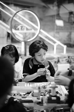 Children are sitting at a table engaged in building with LEGO bricks. The background features a large circular light fixture and a bustling indoor environment, suggesting a playful or educational setting.