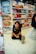 A child with a backpack is sitting on the floor in a store aisle filled with various beauty and personal care products on shelves. Bright colorful packaging and promotional signs accent the background.