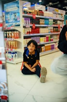 A child with a backpack is sitting on the floor in a store aisle filled with various beauty and personal care products on shelves. Bright colorful packaging and promotional signs accent the background.