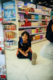 A child with a backpack is sitting on the floor in a store aisle filled with various beauty and personal care products on shelves. Bright colorful packaging and promotional signs accent the background.