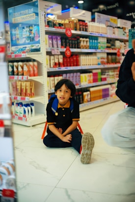 A child with a backpack is sitting on the floor in a store aisle filled with various beauty and personal care products on shelves. Bright colorful packaging and promotional signs accent the background.