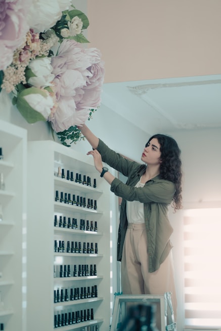 A woman stands on a ladder organizing large, decorative flowers high on a wall. She is positioned next to a shelf filled with organized, small bottles resembling nail polish. The setting appears to be a retail or beauty store, with bright lighting and neat arrangements.