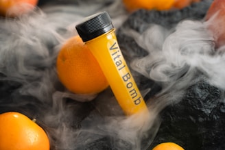 A test tube labeled 'Vital Bomb' is surrounded by oranges, with mist or fog creating a dramatic effect on a dark textured surface.